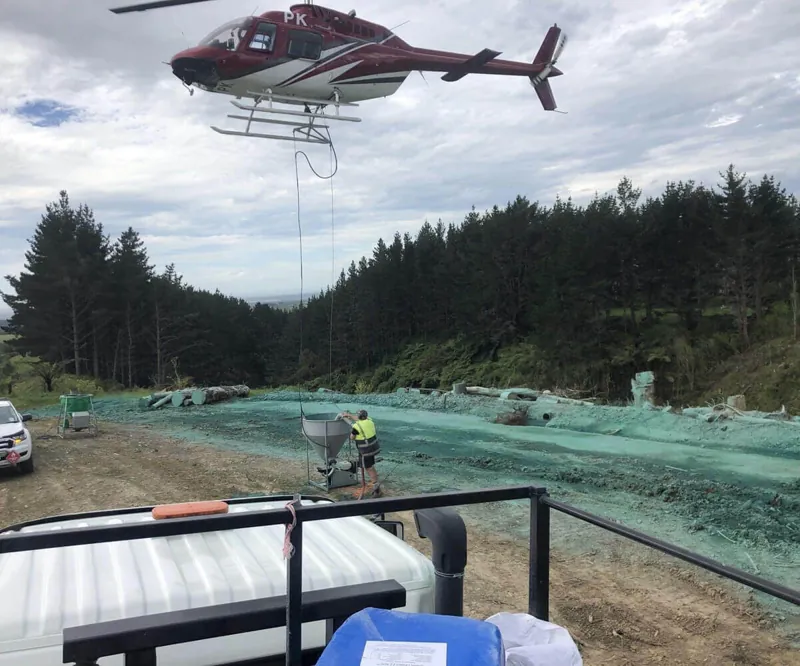 Hubschrauber hebt einen Behälter über einer angespritzten Fläche am Waldrand